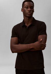 Juodas medvilninis polo marškinėlis su apykakle, trumpomis rankovėmis ir subtiliu logotipu ant krūtinės. Modelis stovi sukryžiavęs rankas.