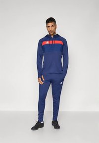 Tengerészkék sportmelegítő vörös vízszintes csíkkal és fehér részletekkel, kapucnis felsővel és szűkített szárú nadrággal. Könnyű anyagból készült.