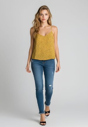 Mujer joven caminando hacia adelante con una camiseta sin mangas amarilla de lunares, vaqueros pitillo azules con rotos y tacones negros de punta abierta sobre un fondo gris.