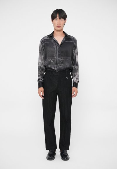 Zwarte zijden blouse met een marmeren patroon, gecombineerd met gestructureerde zwarte broek en zwarte schoenen. De outfit presenteert een monochrome look.