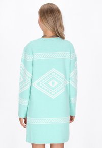 Tīra piparmētru zaļa svētbrīdis ar garām piedurknēm, kas rotāti ar baltām ģeometriskām figūrām un brīvu piegriezumu. Mīksts, teksturēts audums. Vidēja auguma garums.