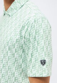 Πουκάμισο με ανοιχτό πράσινο και λευκό καρό σχέδιο, με μικρά λογότυπα Nike και μαύρο έμβλημα Nike με σταυρωτά γκολφ μπαστούνια στο μανίκι.