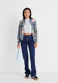 Blomster bomberjakke i blå, cropped grå topp, mørkeblå jeans, svarte joggesko med hvite detaljer, og lyseblå veske. Glatte teksturer.