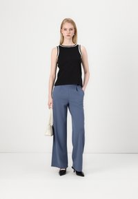 Top senza maniche nero con rifiniture bianche, abbinato a pantaloni larghi azzurri. Il modello tiene una piccola borsa bianca e indossa scarpe con tacco nere.
