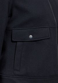 Veste en laine noire avec fermeture à glissière, poche à rabat avec un bouton argenté et texture lisse. Le design est simple et pratique.