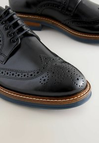 Czarne skórzane buty na Formalne z ostrym noskiem, dekoracyjnymi perforacjami w stylu brogue oraz kontrastującą brązową podeszwą z detalami szewskimi.