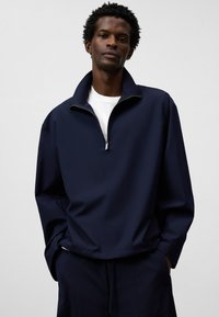 Marinblå halvdraget pullover tillverkad av slät tyg, med en uppstående krage och avslappnad passform. Bärs över en vit T-shirt, med minimala detaljer.