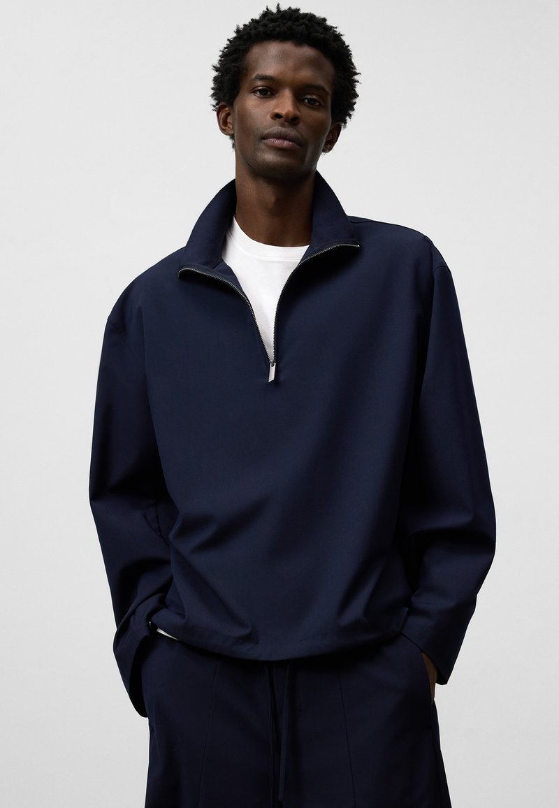 Marinblå halvdraget pullover tillverkad av slät tyg, med en uppstående krage och avslappnad passform. Bärs över en vit T-shirt, med minimala detaljer.