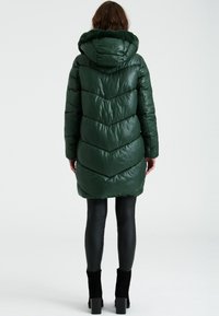 Abrigo acolchado verde con capucha, que presenta un diseño de chevron acolchado. El material parece brillante, con un suave ribete a juego alrededor de la capucha.