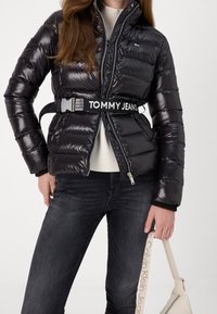 Svart dunjacka med glansig yta, bältad med etiketten "TOMMY JEANS". Bärs över en vit ribbad tröja och matchas med mörka jeans.