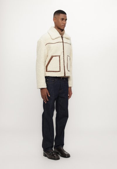 Cream-colored shearling jacket with brown accents, zipper closure, and two front pockets. Worn with dark jeans and black shoes.