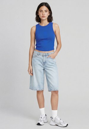 Femme debout portant un crop top bleu sans manches, un short en jean clair jusqu'aux genoux, des chaussettes blanches et des baskets blanches sur un fond gris.