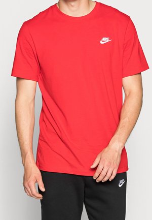 Mężczyzna w czerwonej koszulce Nike i czarnych spodniach Nike, z widocznymi logo na klatce piersiowej i udzie, na jednolitym tle.
