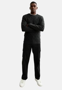 Maglione lavorato a maglia grigio scuro con motivo a diamante; pantaloni cargo neri; sneakers bianche basse; il modello sta in piedi con le braccia incrociate.
