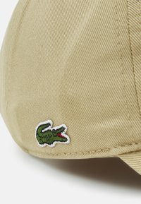 Beige caps laget av teksturert stoff, med en liten grønn krokodillelogo med røde detaljer, plassert på siden av capsen.