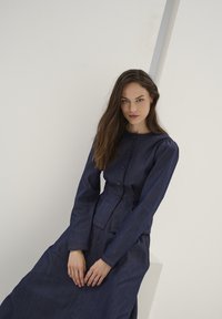 Vestito di jeans blu scuro, con maniche lunghe, scollo tondo e chiusura con bottoni. Dettagli con cuciture visibili e vita aderente.