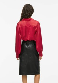 Woman with long curly hair wearing a red satin blouse and a high-waisted black leather pencil skirt, shown from the back.