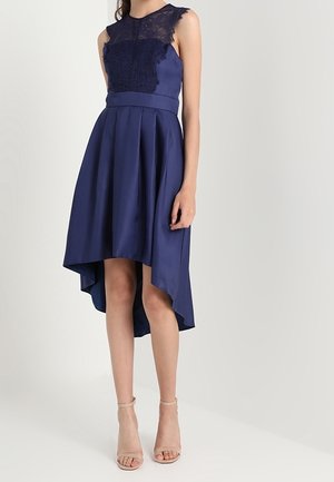 Ærmeløs navyblå kjole med blondeoverdel og høj-lav plisseret nederdel, kombineret med nøgenfarvede ankelrem-sandaler på hvid baggrund.