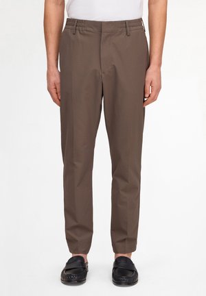 Man wearing brown checkered slim-fit trousers and black leather slip-on loafers, standing against a white background.