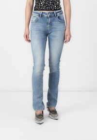 Jean en denim bleu clair à coupe droite, cinq poches et une fermeture classique à bouton et zip. Portés avec des chaussures à lacets métalliques.