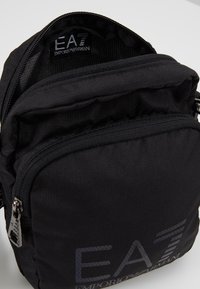 Borsa a tracolla nera in nylon con esterno testurizzato, scomparti con zip e dettaglio del logo. Presenta una chiusura zippata superiore e una tracolla regolabile.