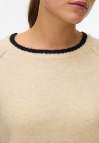 Beige breisel trui met een zwarte geribbelde halslijn, zachte textuur en een ontspannen pasvorm, getoond met een close-up van de kraag.