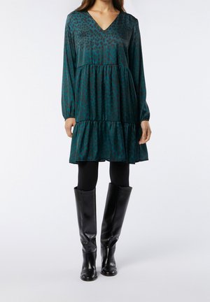 Woman wearing a teal and black leopard print dress, black leggings, and knee-high black leather boots, standing against a plain white background.