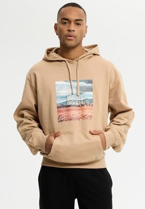 Hombre con sudadera beige con capucha, con las manos en el bolsillo delantero y un estampado de paisaje con la frase "We belong to the Earth" en el pecho sobre un fondo blanco.