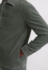 Kurtka sztruksowa w ciemnozielonym kolorze z pionową prążkowaną fakturą, zapinana na guziki, z bocznymi kieszeniami. Posiada kołnierz i elastyczne mankiety.