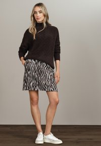 Kvinna som bär en mörkbrun strukturerad tröja, en zebra-mönstrad kjol och vita sneakers, stående på trägolv mot en ljusgrå vägg.