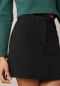 Gros plan sur une personne portant une mini jupe noire taille haute et un haut à manches longues vert foncé, avec une main détendue sur le côté.