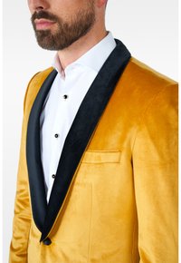 Geltonas aksominis tuxedo švarkas su juodais satino atvartais ir įpjautu kaklu, turintis krūtinės kišenę ir juodus mygtukus ant baltos marškinių.
