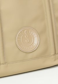 Borsa in tessuto beige con un patch circolare beige con testo in rilievo e cuciture a contrasto lungo i bordi, che mette in risalto una texture liscia.