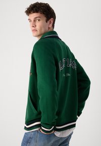 Groene gebreide cardigan met witte en marineblauwe strepen bij de manchetten en onderrand, met geborduurde tekst "MAT 1945" op de achterkant en zijzakken.