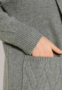 Hand gedeeltelijk in de zak van een grijze gebreide trui met geribbelde en chevrontextuurpatronen op de mouwen en de zak.