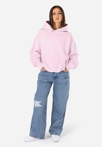 Moletom rosa-claro com bolso frontal e capuz com cordão, combinado com jeans azul largos com um padrão desgastado. Ténis brancos completam o visual.