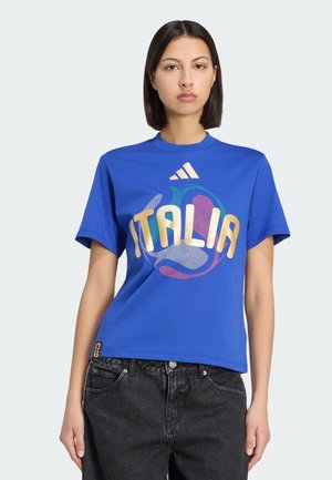 Mujer con cabello lacio y oscuro que lleva una camiseta azul de Adidas con la palabra "ITALIA" y un diseño de globo multicolor, combinada con jeans negros.