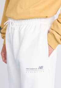 Pantalon de survêtement blanc avec une taille à cordon, comportant des poches latérales et un logo gris "New Balance Athletics" sur la cuisse gauche.