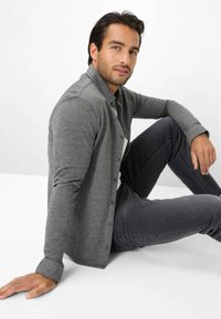 Graues Hemd mit strukturierter Stoffoberfläche, Kragen und langen Ärmeln, kombiniert mit dunkelgrauen Jeans. Modell sitzt auf einer weißen Fläche.