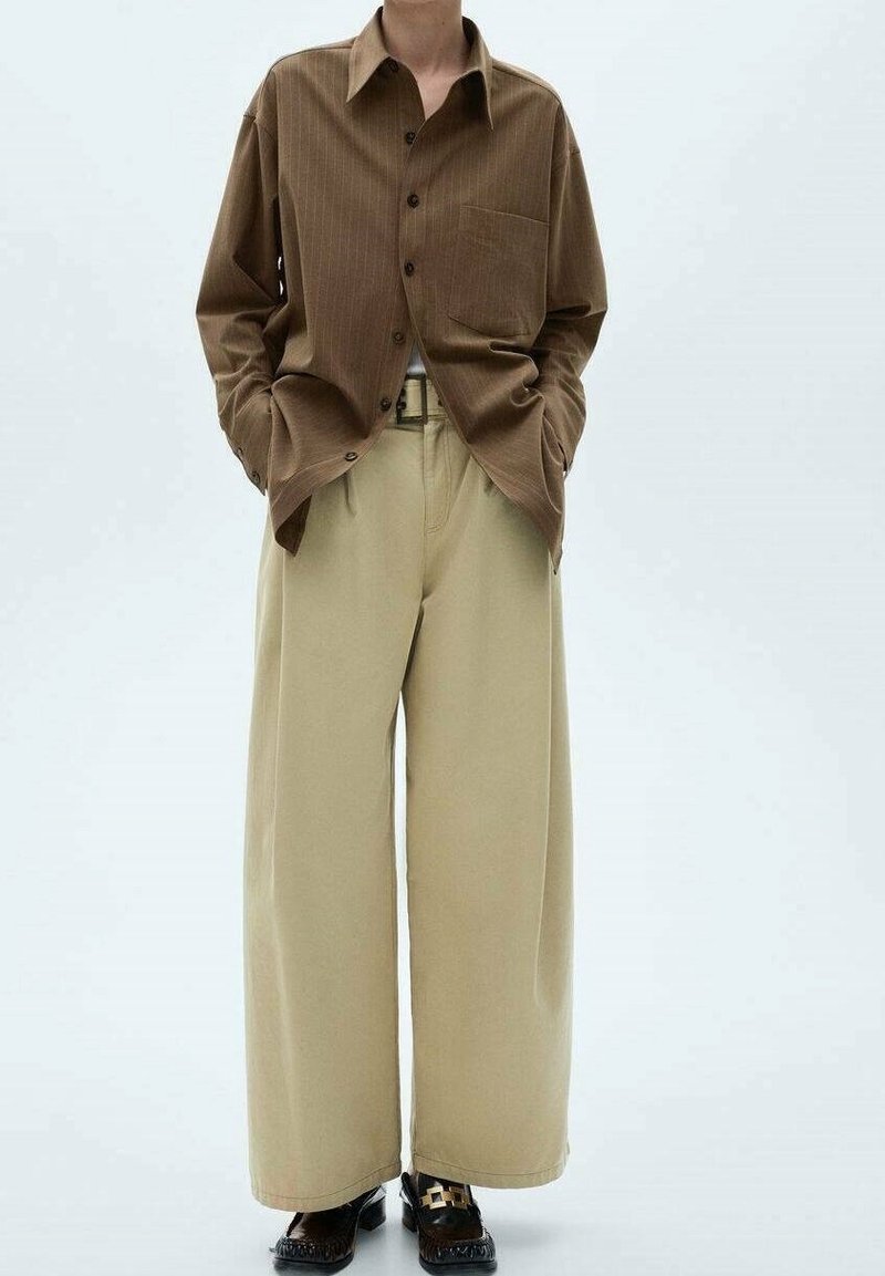 Personne portant une chemise marron à boutons, un pantalon beige à jambes larges, une ceinture noire et des mocassins noirs sur fond blanc. Mains dans les poches.