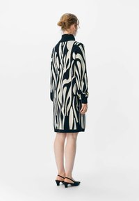 Robe en tricot avec un motif abstrait noir et crème, col montant, manches longues et une longueur aux genoux, accompagnée de talons slingback noirs.