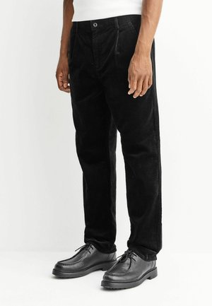 Schwarze Cordhosen mit geradem Bein, ausgestattet mit einem Knopfverschluss und vorderen Falten. Kombiniert mit schwarzen Lederschuhen.