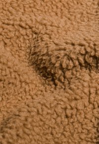 Brun texturerad tyg med en mjuk, fluffig yta som har ojämna, upphöjda mönster över hela. Materialet verkar varmt och mysigt.