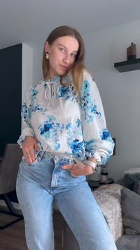 Blusa floral com padrões azuis e aquáticos, tecido texturizado, punhos franzidos e um laço no pescoço. Combinada com jeans de cintura alta em denim claro.