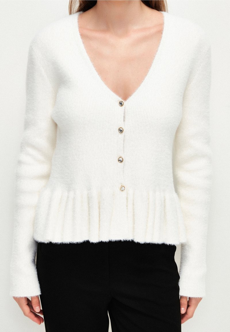 Femme portant un cardigan blanc duveteux à col en V avec quatre boutons à l'avant et un bas peplum, associé à un pantalon noir.