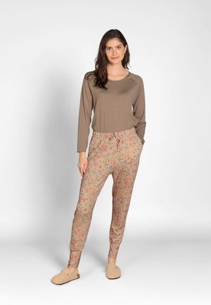 Frau steht, trägt ein braun gestreiftes Langarmshirt, beige Hose mit Blumenmuster und beige flauschige Hausschuhe vor weißem Hintergrund.