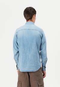 Lichtblauw denim shirt met lange mouwen, knoopmanchetten en een kraag. Heeft een rechte snit en een rugnaadontwerp. Zichtbare stikseldetails.