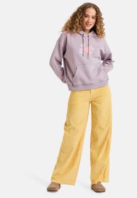 Levanduľová mikina s kapucňou a kvetinovým vzorom, v kombinácii so žltými širokými nohavicami z kordu. Outfit dopĺňajú béžové topánky.