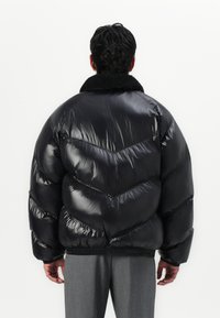 Zwarte pufferjack met een glanzende, soepele textuur, voorzien van een chevronpatroon en een zachte, met zwart bont gevoerde kraag. Elastische manchetten en zoom.