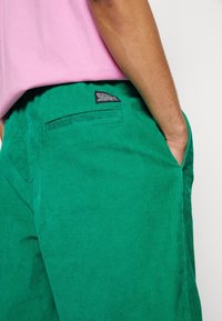 Gröna kordfläskbyxor med texturerad yta, sidofickor och en bakficka med ett märkt etikett. Bärs med en rosa t-shirt.